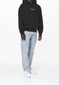 Muž v černé mikině "PEGADOR", světle modrých džínách a bílých teniskách stojí na pozadí z jedné barvy - bílé.