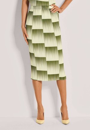 Vrouw draagt een geplooide midi-rok met een groen verlopen dambordpatroon en gele hoge hakken, staand met één hand op de heup.