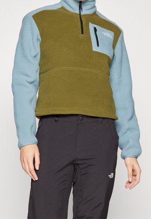Persoon draagt een groen en lichtblauw fleece pullover met zwarte ritszak en zwarte outdoorbroek met The North Face-logo.