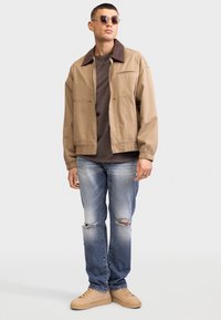 Giacca marrone chiaro con colletto scuro, t-shirt marrone, jeans blu strappati e sneakers beige. Occhiali da sole rotondi completano l'outfit.