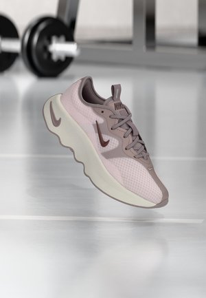 Hellvioletter Nike-Laufschuh mit dicker weißer Sohle, der in einem Innenraum auf dem Hallenboden schwebt, im Hintergrund verschwommene Gewichthebergeräte.