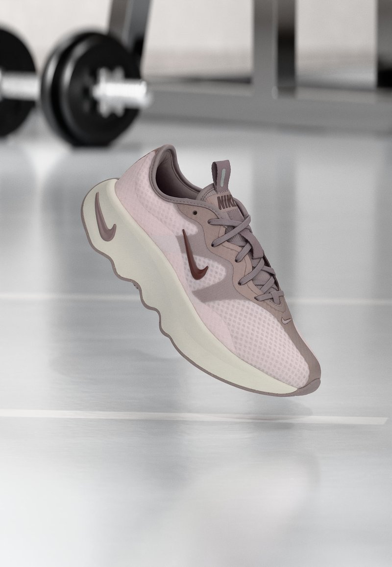 Zapatilla de correr Nike color púrpura claro con suela blanca gruesa, flotando en el interior sobre el suelo de un gimnasio con equipo de levantamiento de pesas desenfocado en el fondo.