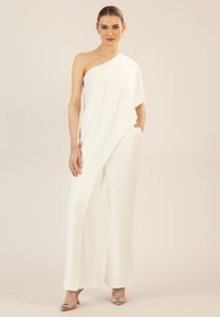 Combinaison blanche à épaule unique avec un design drapé, jambes larges et poches latérales. Tissu lisse, style minimaliste, assortie à des talons ornés.