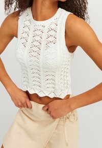 Vit stickad ärmlös topp med ett strukturerat mönster, scalloped nederkant och en figurnära design. Bärs med beige shorts med knytning i midjan.