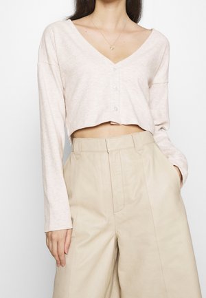 Woman wearing a light pink cropped buttoned cardigan and high-waisted beige wide-leg trousers, with one hand in pocket.