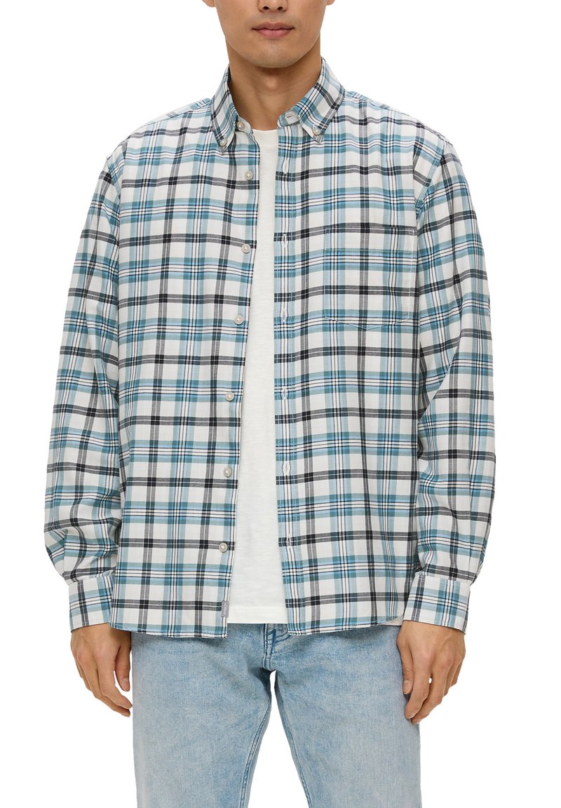 Homme portant une chemise à carreaux bleue, noire et blanche par-dessus un t-shirt blanc avec un jean bleu clair.