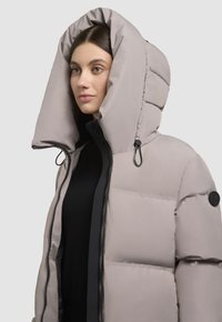 Chaqueta acolchada gris claro con capucha oversized, cierre de cremallera y acentos negros. Presenta un patrón de burbujas texturizado y un parche con el logo en el hombro.