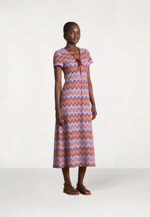 Femme debout portant une robe midi à manches courtes et col en V avec un motif zigzag multicolore, ainsi que des sandales plates marron à brides autour de la cheville.