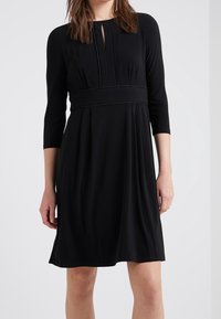 Musta polvipituinen mekko pehmeästä jersey-kankaasta, jossa on pyöreä pääntie avaimenrei'ellä ja kolmanneksen pituiset hihat, kerätty vyötärö.