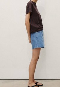 Personne portant une chemise marron foncé à manches courtes, un short en jean bleu clair et des sandales noires, debout de profil contre un mur uni.