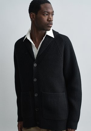 Cardigan nero a coste con collo a scialle, chiusura a bottoni e due tasche frontali. Il tessuto texturizzato conferisce profondità al design complessivo.
