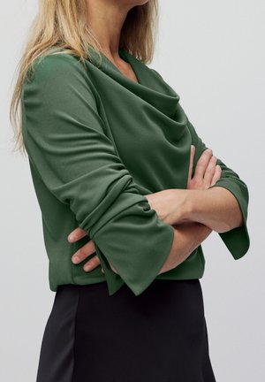 Mujer con cabello largo y rubio lleva una blusa verde de manga larga con escote drapeado y falda negra, con los brazos cruzados frente a un fondo liso.