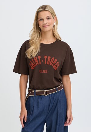 Femme aux longs cheveux blonds portant un t-shirt marron Saint-Tropez Club, un pantalon bleu avec une ceinture, debout devant un fond clair uni.