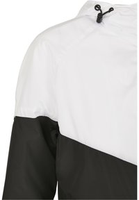 Vindjacka med vit övre del och svart nedre del, med en slät yta och justerbar huva.