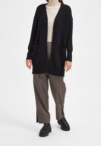 Svart lång cardigan med öppen framsidan, två framfickor och lös passform. Bärs över en krämfärgad turtleneck, tillsammans med bruna rutiga byxor och svarta stövlar.