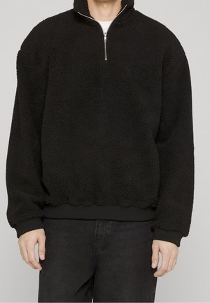 Mężczyzna noszący czarny, teksturowany polarowy sweter z kołnierzem na zamek do ćwiartki, ściągaczami na mankietach i u dołu, w połączeniu z czarnymi spodniami.