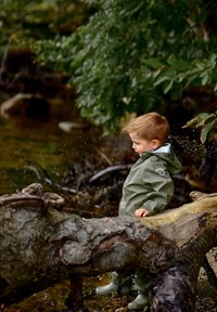 Niño con chaqueta impermeable verde claro y botas a juego, de pie sobre un tronco cerca del agua, rodeado de árboles y gotas salpicando.
