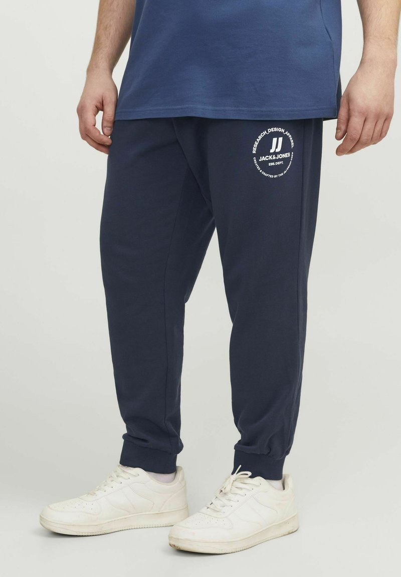 Marina byxor med mudd, med en cirkulär vit logotyp "JACK & JONES" på låret. Matchas med vita sneakers.