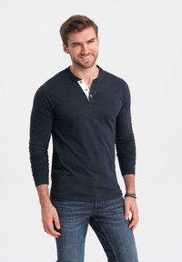 Marineblaue Langarm-Henley-Shirt mit einem kontrastierenden hellen Kragen und drei Knöpfen. Kombiniert mit blauen Jeans. Aus glattem Baumwollmaterial gefertigt.