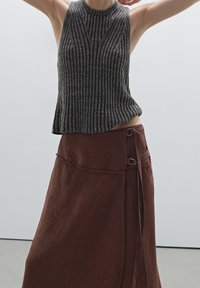 Nainen, jolla on yllään hihaton harmaa ribbeknitti toppi ja korkeavyötäröinen ruskea kietaisuhame, jossa on solkilukit, poseeraa käsivarsia nostettuna.