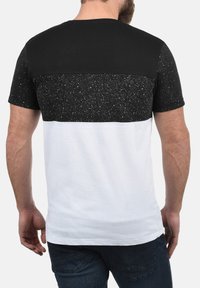 Una camiseta de manga corta en blanco y negro con un diseño moteado. Presenta una sección superior completamente negra y una sección inferior blanca. Tejido de algodón suave.