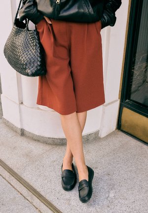 Persona che indossa pantaloncini color ruggine fino al ginocchio, mocassini neri e una giacca di pelle nera, tenendo una borsa nera intrecciata.