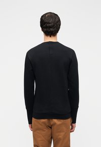 Μαύρο φούτερ με μακριά μανίκια, λαιμόκοψη σε στρογγυλή γραμμή, υφασμάτινη υφή και λεπτομέρεια με κεντρικό ραφή στο πίσω μέρος. Συνδυάζεται με μπεζ παντελόνι.