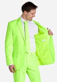 Neongrüner Anzug mit zwei Knöpfen, passender Krawatte und weißem Hemd. Die Jacke ist offen, wodurch ein schlichter Innenraum mit einem Etikett sichtbar wird.