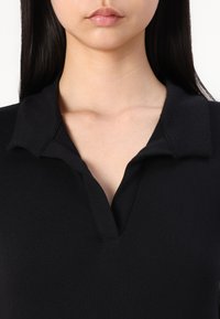 Polo noir en tissu coton texturé, avec un col et un décolleté en V distinct. Le design est minimal et ajusté.