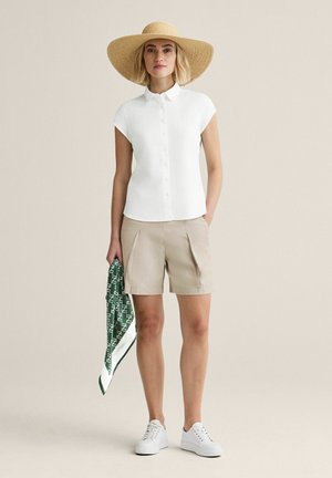 Nainen pukeutunut valkoiseen lyhythihaiseen nappipaitaan, beigen värisiin shortseihin, valkoisiin tennareihin, leveälieriseen olkihattuun ja pitää kädessään vihreää kuviollista huivia.