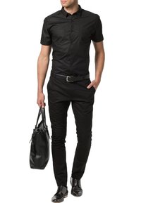 Camisa negra de manga corta con dos bolsillos frontales, combinada con pantalones negros y un bolso de cuero a juego. Zapatos negros brillantes completan el look.