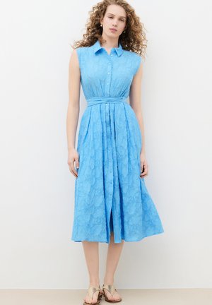 Femme aux cheveux bouclés portant une robe midi en dentelle bleue sans manches avec des boutons, un col et une ceinture nouée à la taille, debout contre un mur blanc.