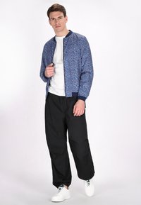 Blauwe gebreide bomberjack met een rits, geribde manchetten en zoom; gecombineerd met losse zwarte broek en witte sneakers.