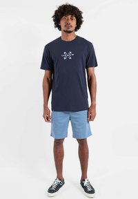 T-shirt en coton bleu marine avec un motif central bleu clair, associé à un short bleu clair. Le modèle porte des baskets noires avec des lacets blancs.