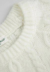 Biały sweter dzianinowy z prążkowanym kołnierzem i wzorami w warkocz. Materiał wydaje się miękki i puszysty, co podkreśla jego teksturowany wygląd.