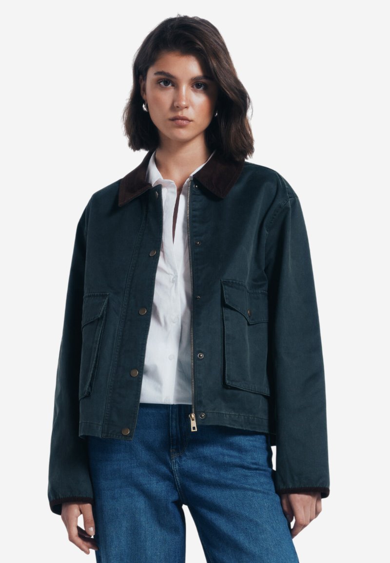 Mörkgrön jacka med brun krage, knappstängning fram och två överdimensionerade fickor. Bärs över en vit skjorta med långa ärmar och avslappnad passform.