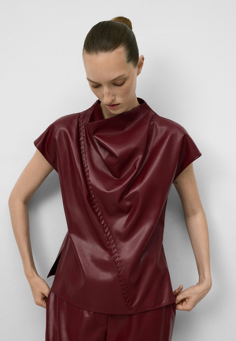 Top in ecopelle bordeaux lucida con scollatura drappeggiata, maniche corte e dettagli con treccia laterale. Texture liscia, design aderente e silhouette contemporanea.