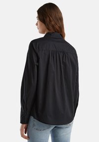 Maglietta nera a maniche lunghe con colletto classico, caratterizzata da un dettaglio arricciato sul retro e polsini bottone, realizzata in un tessuto liscio e intrecciato.