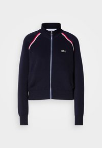 Veste bleu marine zippée avec col et poignets côtelés, rayures rouges et blanches sur les épaules, et petit logo crocodile vert sur la poitrine.