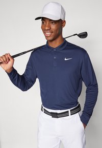Marinblå långärmad polo med knäppt krage och vit Nike-logotyp, kombinerat med vita byxor och ett svartrandigt bälte. Håller en golfklubba.