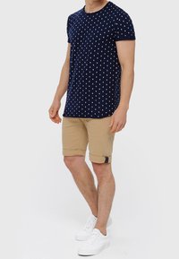 Camisa de manga corta azul marino con lunares blancos, combinada con pantalones cortos beige con puños doblados y un acento negro. Llevado con zapatillas blancas.