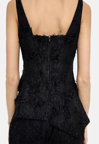 Czarna sukienka o teksturowanej powierzchni, z dekoltem w kształcie łódki, warstwowym materiałem z postrzępionymi brzegami oraz centralnym zapięciem na zamek.