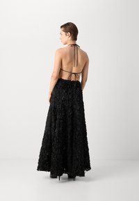 Robe maxi noire à fleurs avec un col halter et un dos nu. Elle présente un tissu texturé avec des motifs floraux superposés et des liens au niveau du cou.