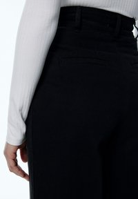 Pantalones de mezclilla de talle alto negros, con bolsillos traseros. La tela tiene una textura suave y se muestra junto a una blusa de manga larga blanca de canalé.