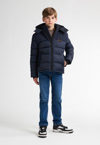 Jachetă puffer navy cu fermoar în față, glugă ajustabilă și buzunare laterale, purtată cu blugi albaștri și adidași negri și albi.
