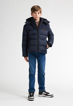 Chaqueta acolchada azul marino con cremallera frontal, capucha ajustable y bolsillos laterales, combinada con jeans azules y zapatillas deportivas negro/blanco.
