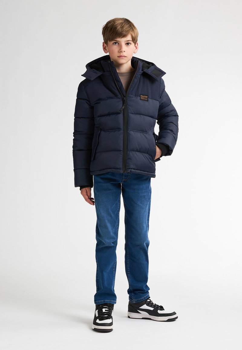 Jachetă puffer navy cu fermoar în față, glugă ajustabilă și buzunare laterale, purtată cu blugi albaștri și adidași negri și albi.