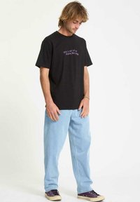 Camiseta de algodón negra con texto en blanco y púrpura, combinada con pantalones de mezclilla azul claro. El corte es holgado, con mangas cortas y un escote redondo.