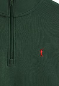 Pullover verde con zip realizzato in un morbido tessuto. Presenta una piccola figura ricamata rossa con un bastone da golf sul petto sinistro. Finitura testurizzata.