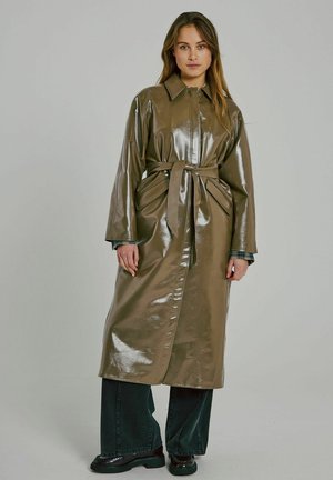 Lång olivgrön trenchcoat gjord av glänsande, strukturerat material med knytmidja och stora, draperade fickor. Matchad med mörka vida byxor.
